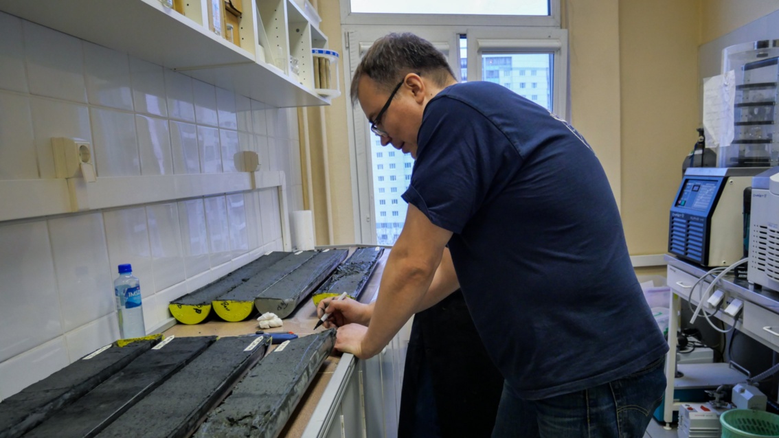In einen Labor liegen mehrere regenrinnenartige, mit Schlamm gefüllte Gefäße auf einer Arbeitsplatte. Ein Mann schaut sich deren Inhalt an.