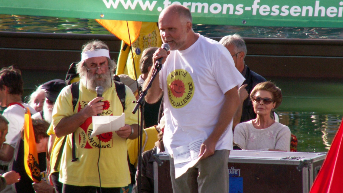 Auf einer Bühne stehen zwei ältere Herren, jeweils mit Mikrofonen in der Hand. Beide tragen T-Shirts mit der Anti-Atom-Sonne.