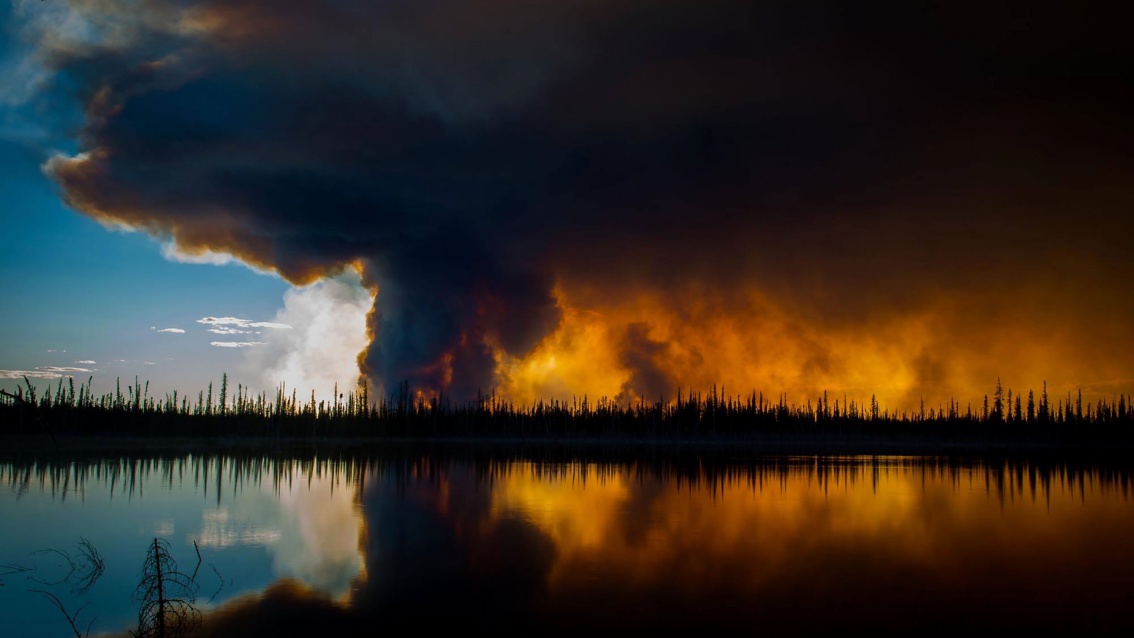 Eine riesige Feuerwalze frisst sich durch einen dürren Wald, im Vordergrund spiegelt sich das Geschehen im Wasser eines Sees