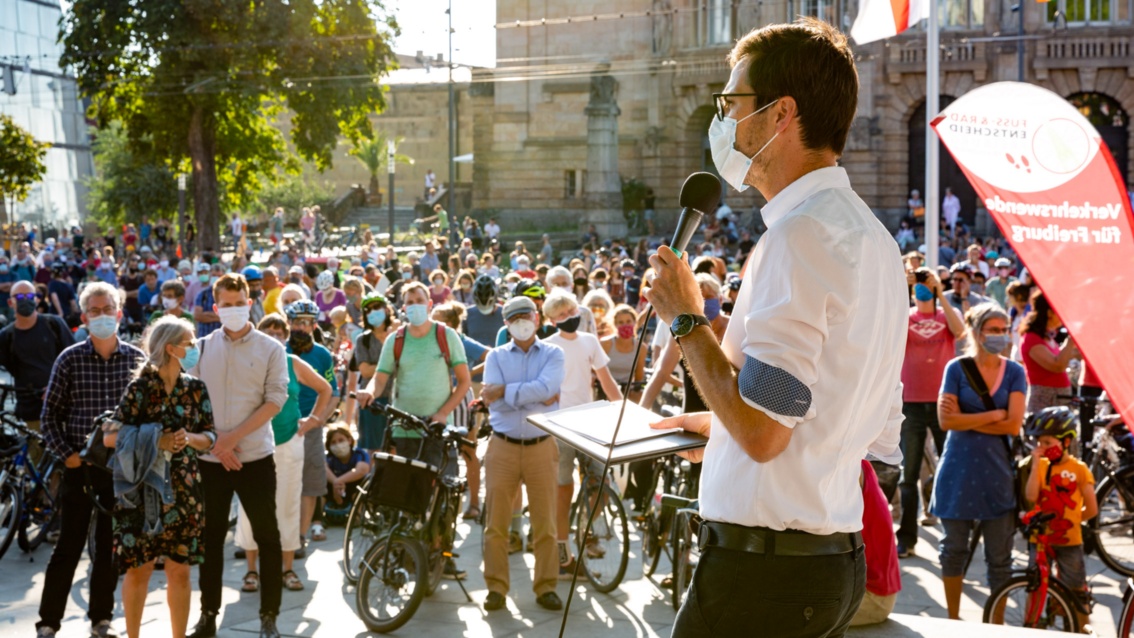 Auf einem sommerlichen städtischen Platz spricht ein Mann vor vielen Menschen mit Fahrrädern.