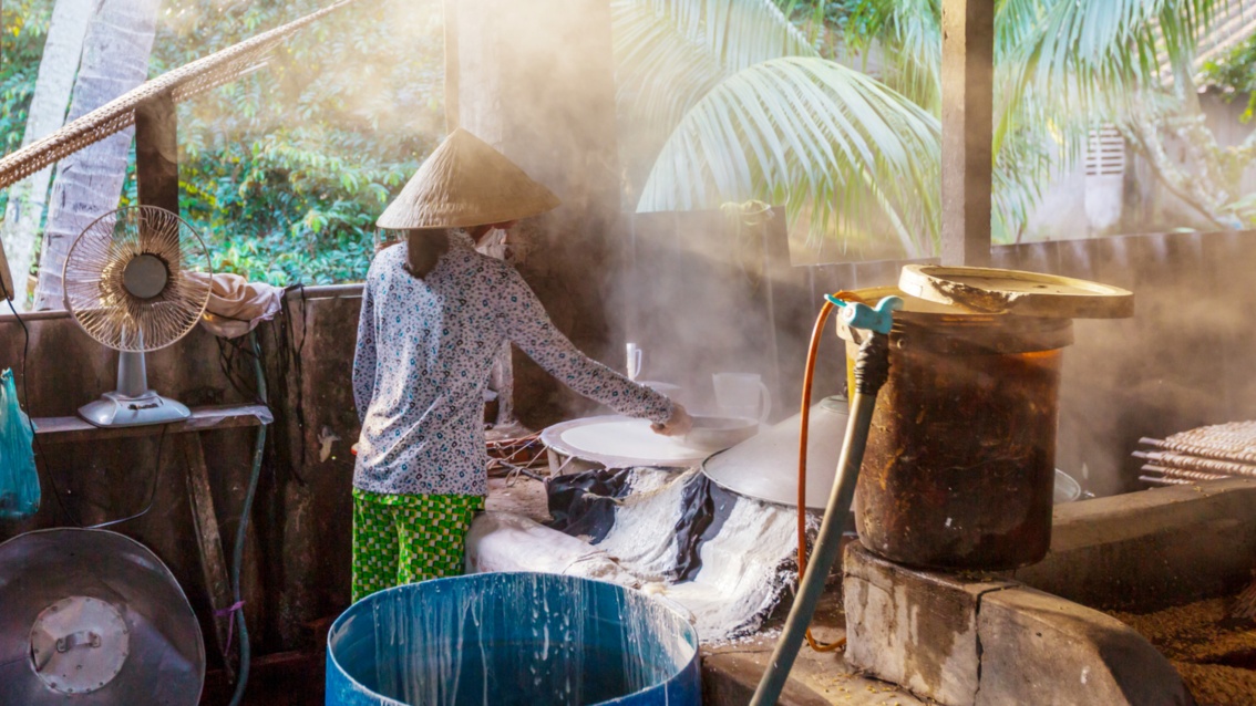 In einer offenen Küche steht eine Frau mit einem Bambushut vor einem dampfenden Kessel.