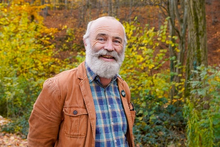 In einem herbstlich bunten Wald steht ein älterer grauhaariger Mann mit einem Vollbart und schaut offen lächelnd in die Kamera.