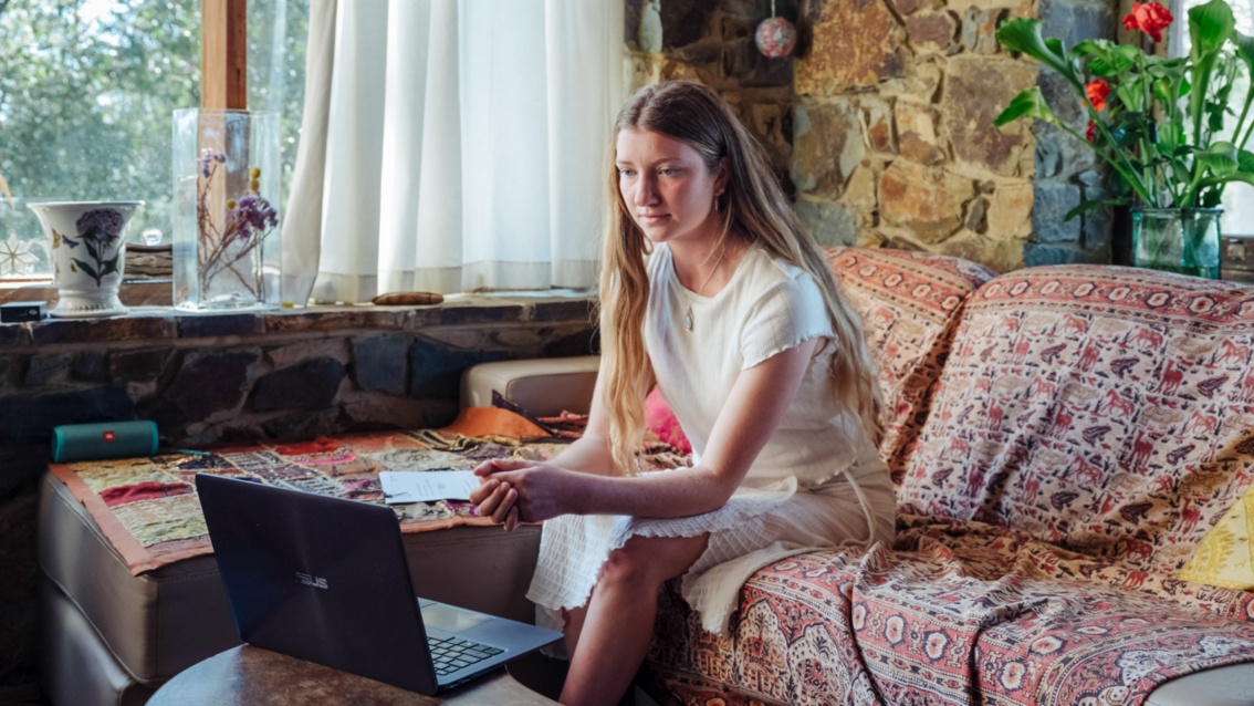Eine junge Frau sitzt in sommerlicher Kleidung auf einem Sofa und schaut in ihren Laptop.