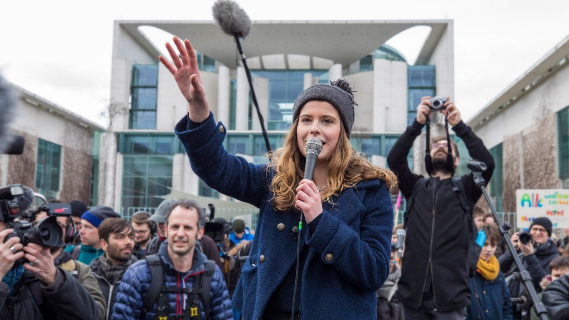 Eine junge Frau steht vor dem Bundeskanzleramt und hält offensichtlich eine Rede, umringt von Menschen, Kameras und Mikrofonen.