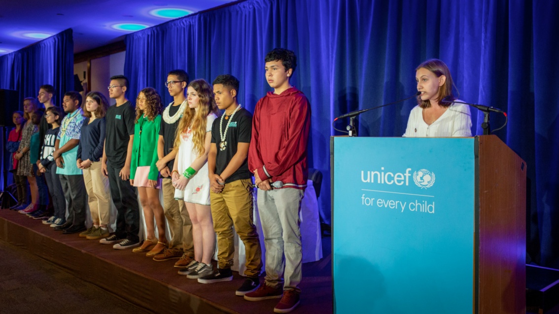 Eine junge Frau hält ein einem Pult mit der Aufschrift UNICEF eine Rede. Daneben stehen etwa zehn  junge Menschen in einer Reihe auf der Bühne.
