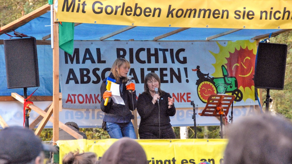 Auf einer zusammengezimmerten Bühne mit gelben Anti-Atom-Transparenten stehen zwei Frauen mit Mikrifonen und singen.