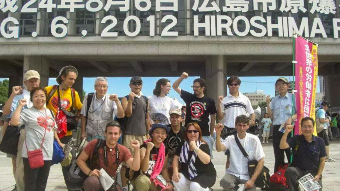 Eine große Menschengruppe vor einem Gebäude, auf dem große japanische Schriftzeichen prangen und in lateinischen Buchstaben «Hiroshima» steht.