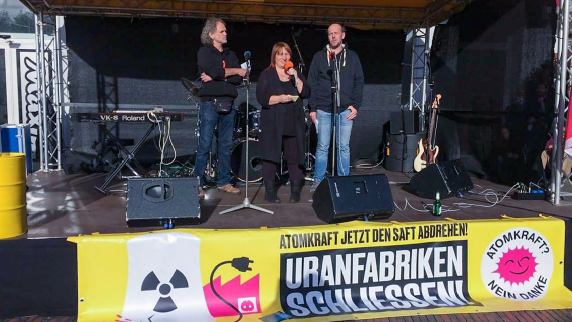 Vor einer Demo-Bühne hängen Transparente mit dem Aufdruck «Uranfabriken schließen». Auf der Bühne, umrahmt von zwei Männern, spricht eine Frau ins Mikrofon.