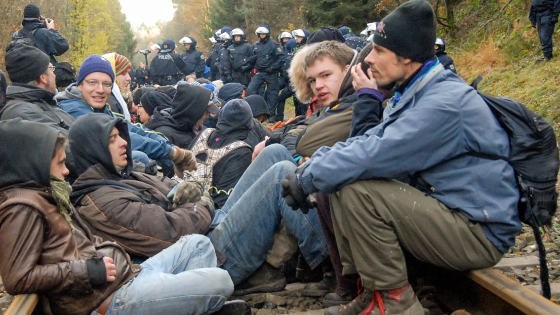 Auf Bahnschienen sitzen sich eine Handvoll junger Menschen in warmer Kleidung gegenüber, im Hintergrund sind zahlreiche Polizisten in Kampfmontur zu sehen. 