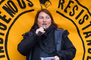 Vor einem großen gelben Transparent spricht eine Frau in ein Mikrofon.