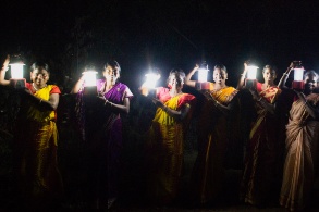 In der Dunkelheit steht eine Reihe indischer Frauen und Männer. Jede hält eine strahlende Lampe in die Höhe. 