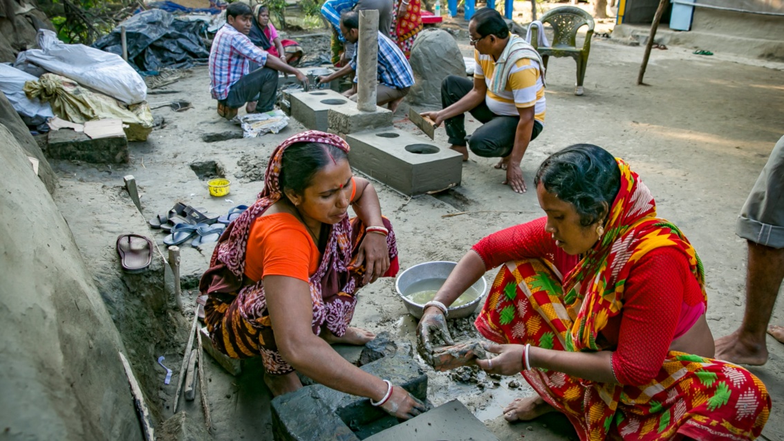 Zwei Frauen in bunten Saries sitzen am Boden über einen aus Lehm hergestellten Ofen-Rohling.