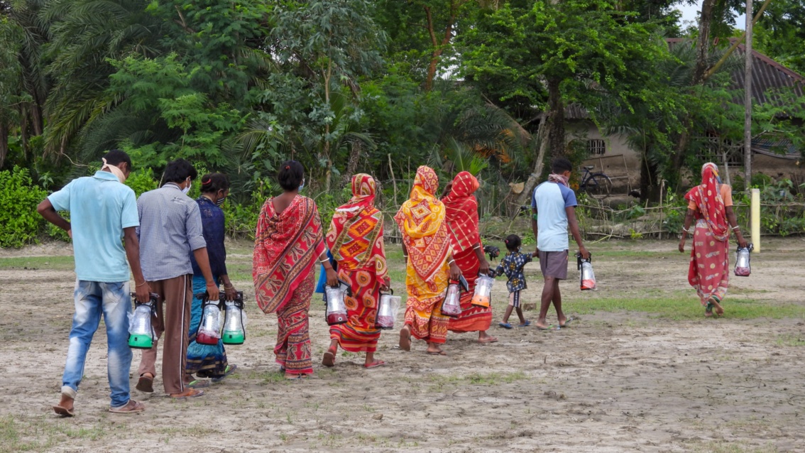 In ländlicher Umgebung entfernt sich eine Reihe in Saris gekleideter Frauen und Männer. Jeder trägt eine Lampe in der Hand.