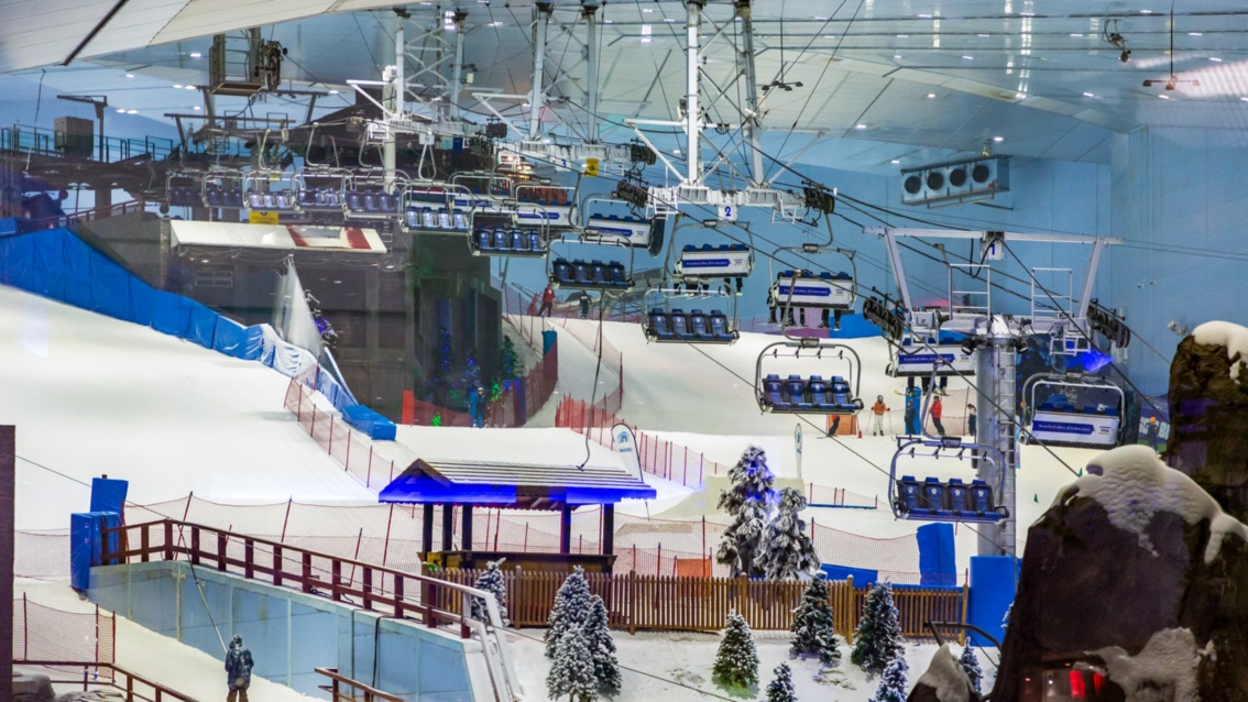 In einer Halle sieht man eine modellhafte Schnee-Landschaft mit Skilift, Skipiste und Almhütte. Von der Decke hängen riesige Klimaanlagen.