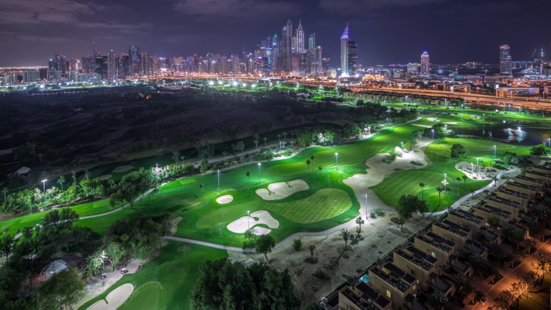 Blick auf eine riesige Golfanlage, die ein der Nacht beleuchtet ist, im Hintergrund die hellerleuchtete Stadt Dubai