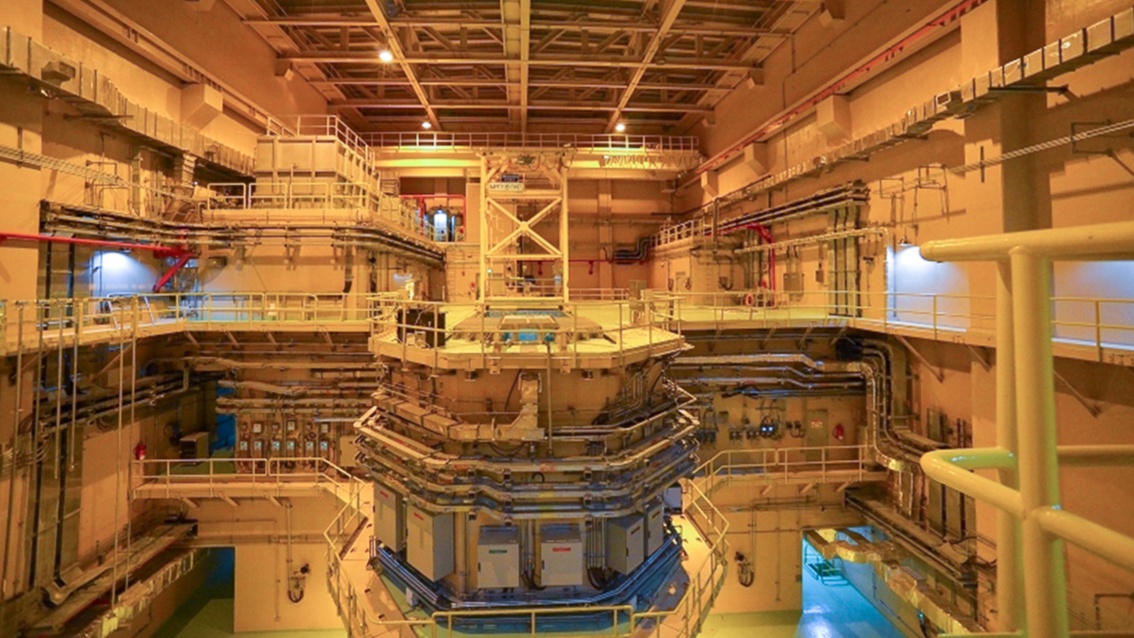 Blick in das Innere eines Reaktorgebäudes mit mehreren Etagen voller Rohre und technischer Anlagen; alles in gelbes Licht getaucht
