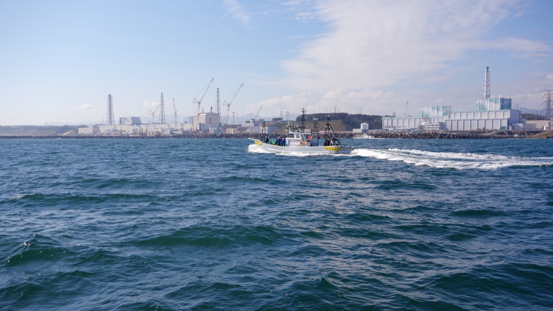Vom Meer aus seiht man ein kleines Boot, an der Küste die Blöcke des Atomkraftwerkes Fukushima.