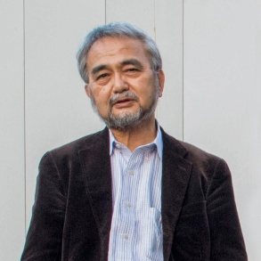 Herr Doi, ein Mann in mittleren Jahren, mit grauem Baart und grauen Haaren steht vor einer weißen Wand.