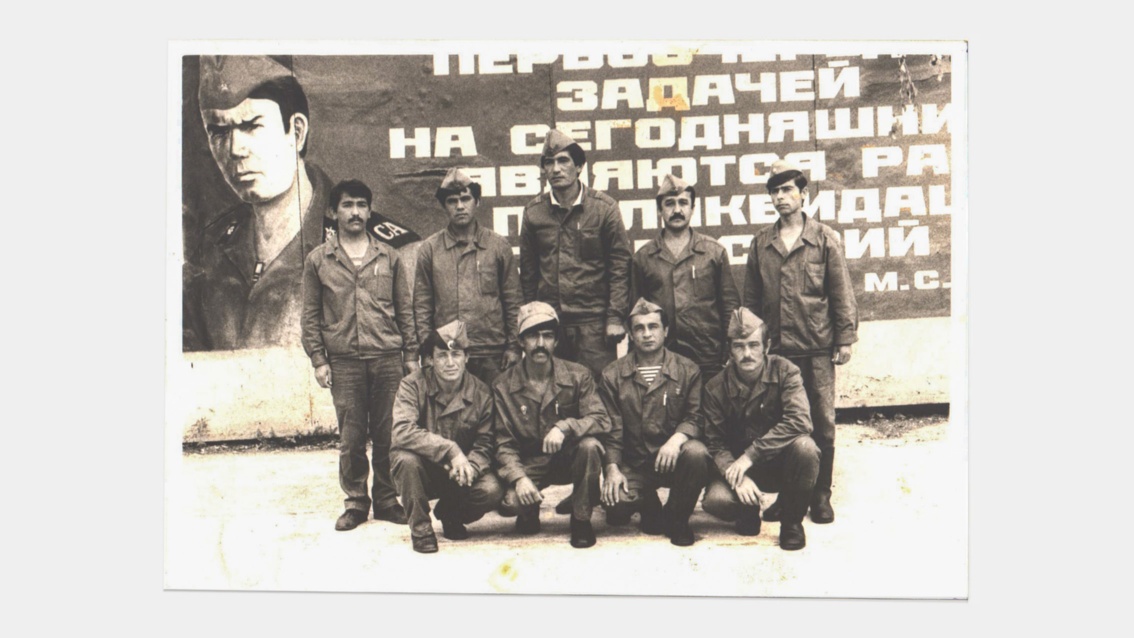 Vor einem großen Propagandaplakt hat sich eine Gruppe von ungefähr 10 jungen Männern zum Gruppenbild aufgestellt.