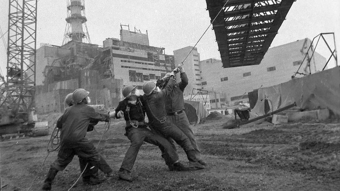 Eine Gruppe Männer zieht vor der Ruine des AKW Tschernobiyl an einem Seil, das an einem über ihnen schwebenden Stahlträger befestigt ist.