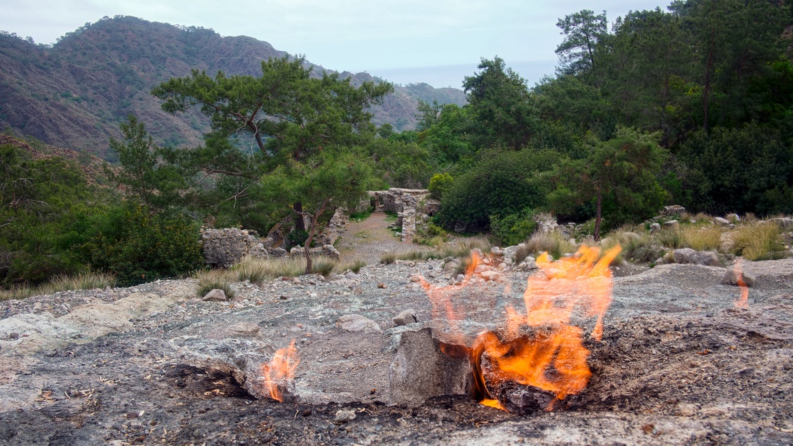 Aus einem steinigen Untergrund vor einer hügeligen Waldlandscheft schlagen große Flammen aus dem Boden.