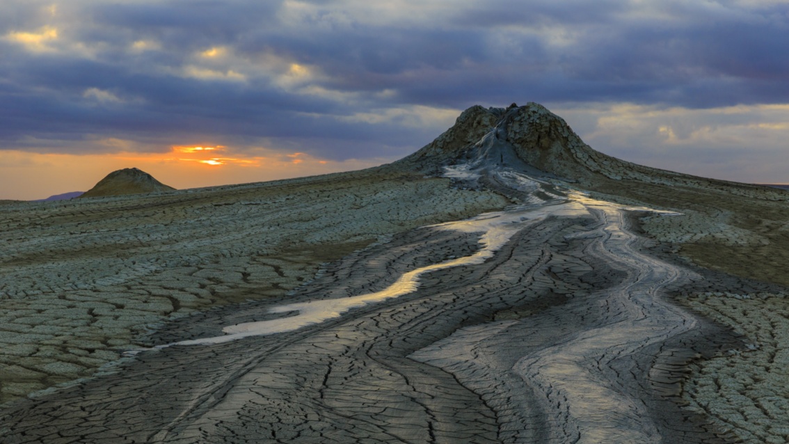 Beeindruckendes Bild einer Schlammlandschaft, im Hintergrund erhebt sich ein Vulkan vor einem Sonnenuntergangshimmel.