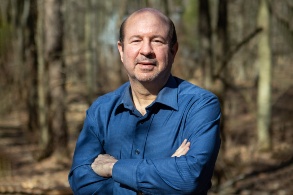 Ein Mann mit schütterem Haar und im blauen Hemd steht mit verschränkten Armen im Wald und blickt ernst in die Kamera.