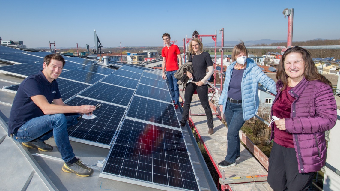 Auf einem Dach das mit Solarpanelen bestückt ist, steht eine Gruppe von Leuten.