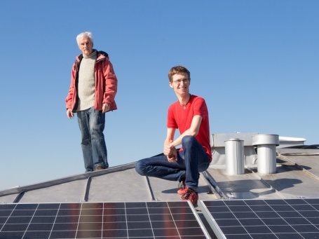 Auf dem Dach sitzt ein junger Mann vor Solarpanelen in der Hocke und ein älterer Herr im Hintergrund.