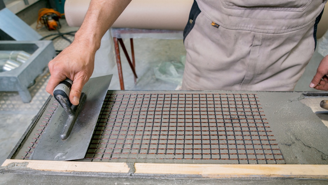 Die Hand eines Arbeiters führt eine große Maurerkelle über ein Stück Beton und streicht so eine Matte in den frischen Beton ein.