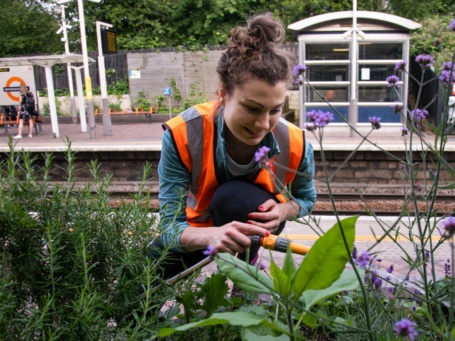 Eine junge Frau in orangefarbener Warnweste gießt Pflanzen etwas oberhalb eines Bahnsteiges der Londoner U-Bahn.