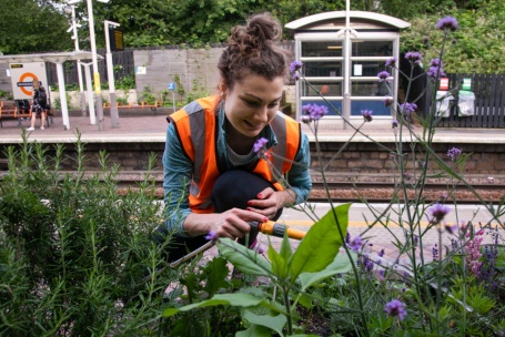 Eine junge Frau in orangefarbener Warnweste gießt Pflanzen etwas oberhalb eines Bahnsteiges der Londoner U-Bahn.