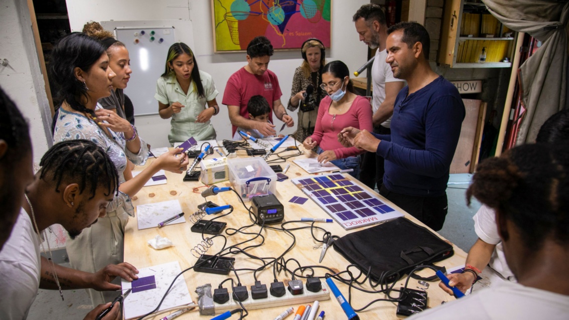 Um einen Werkstatttisch – gefüllt mit kleinen Solarpanelen, Lötkolben und anderem Werkzeug – stehen zehn junge Leute und werkeln.