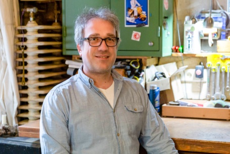 In einer Kellerwerkstatt sitzt ein Mann mit grauen Haaren in Jeans an einem aufgeräumten Werktisch und schaut in die Kamera.