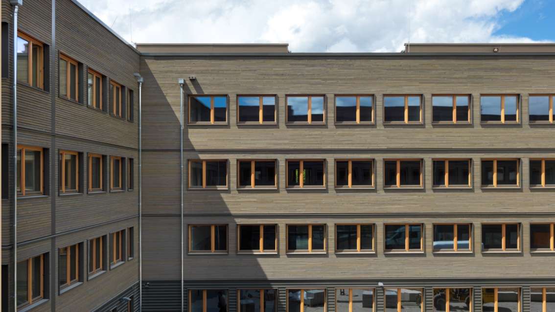 Zwei rechtwinklig zueinander angeordnete Gebäudeflügel, die Fassade ist graubraun, die etwas zurückspengenden Fensteröffnungen sind hellbraun ausgeführt.