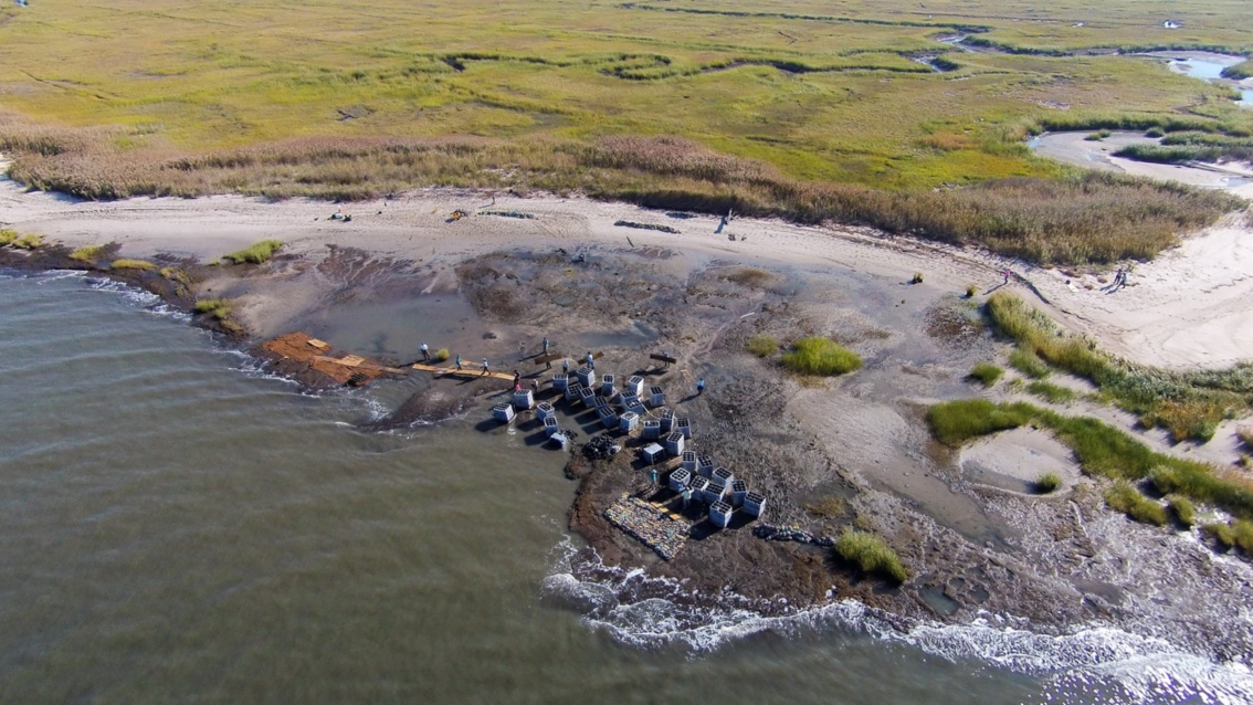 Luftaufnahme einer Küstenlandschaft. An einem Strandstück liegen zahlreiche Betonkästen.