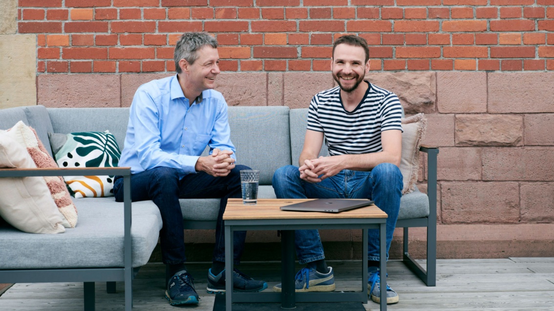 Zwei Männer sitzen in einer Gesprächssituation auf einem Sofa, einer von ihnen schaut lachend in die Kamera.