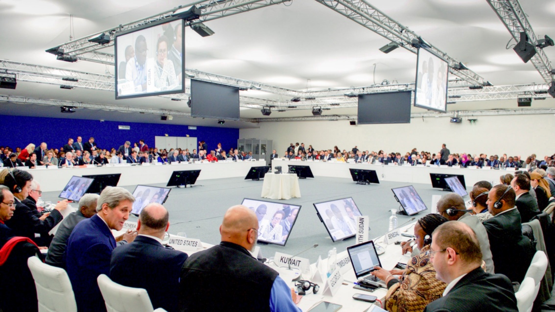 In einem sehr großen Sitzungssaal sitzen mehr als einhundert Delegierte an einem quadratischen Konferenztisch.
