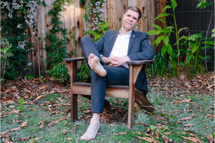 Ein großer Mann, gekleidet im blauen Anzug, sitzt mit nackten Füßen in einem Gartenstuhl vor einem Holzschuppen.