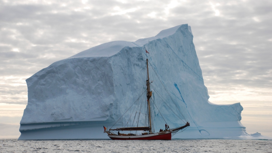Ein riesiger kubischer Eisberg ragt aus dem Wasser, davor liegt das zerbrechlich wirkende Segelschiff mit eingeholten Segeln.