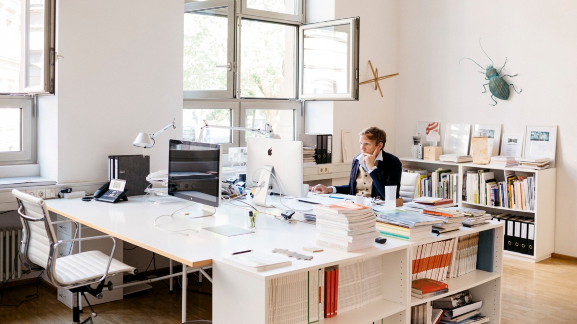 In. einem hellen, sehr großzügigen Büroraum sitzt ein Mann an einem weißen Schreibtisch, im Hintergrund hängt ein großer bronzener Käfer an der Wand.