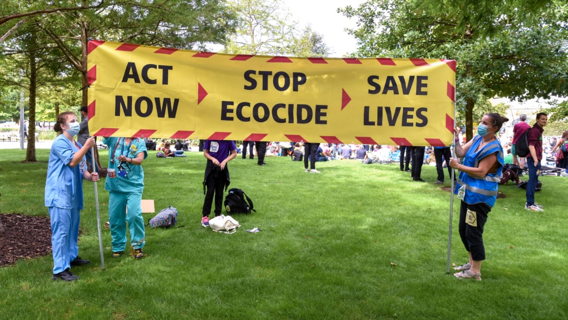 Eine Demo in einem Park: zwei Personen halten ein großes gelbes Transparent mit der Aufschrift: Jetzt Handel, Stop Ökozid, Leben retten.