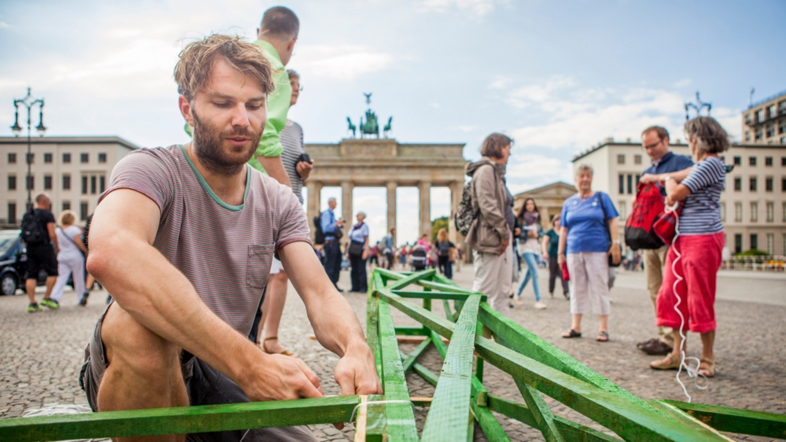 A young man kneels on the ground in front of the Brandenburg Gate, nailing together green wooden slats.