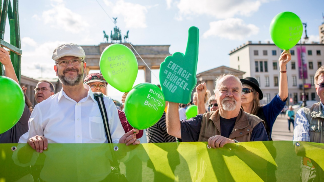 Several people stand behind a green banner in front of the Brandenburg Gate, holding up green foam hands and green balloons.
