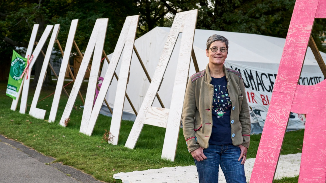 Eine Frau mit grauem Kurzhaarschnitt steht neben menschengroßen Buchstaben, die das Wort Klima bilden.