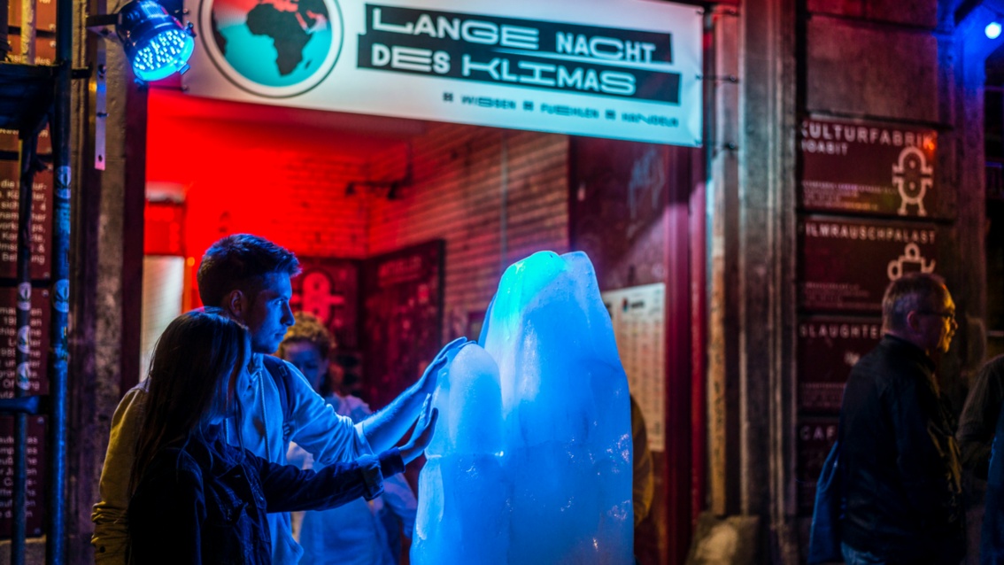 Vor dem bunt beleuchteten Eingang zu einer Veranstaltung taut ein mannshoher Eisblock. Ein junges Paar betastet fasziniert die kalte Oberfläche.