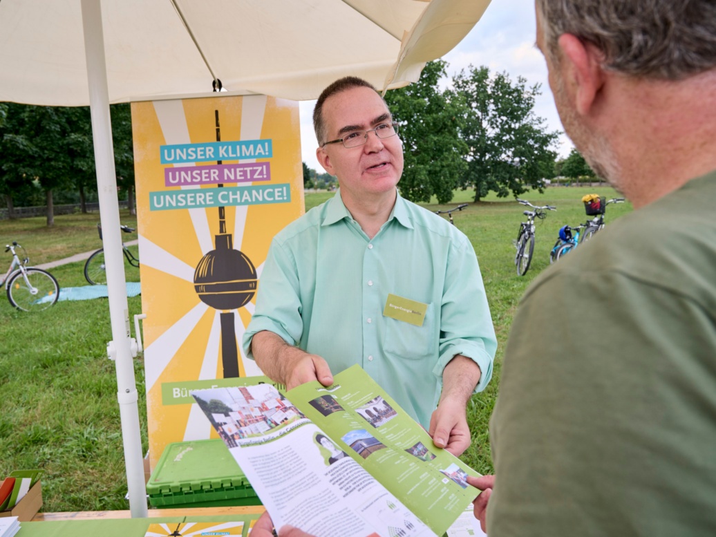 At an information stand set up on a green, summery meadow, a friendly middle-aged man offers a brochure to a man in the foreground.