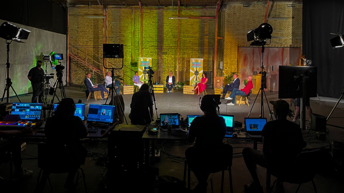 In einer Industriehalle ist ein Fernsehstudio mit vielen Kameras und Monitoren aufgebaut. Auf der Bühne sitzt im Halbrund ein Diskussionsrunde.