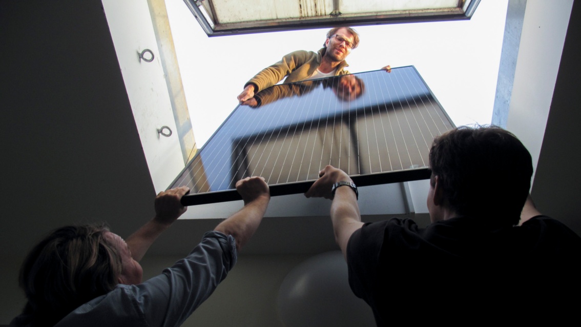 Two men pass a PV module through a skylight to a young man on the roof.