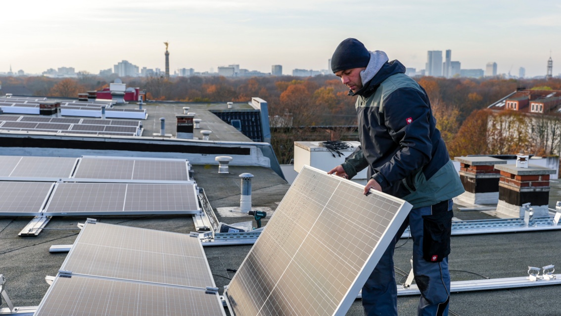 Auf dem Dach eines Hauses steht ein Handwerker und installiert ein Photovoltaik-Panel. Im Hintergrund ist die Silhouette von Berlin erkennbar.